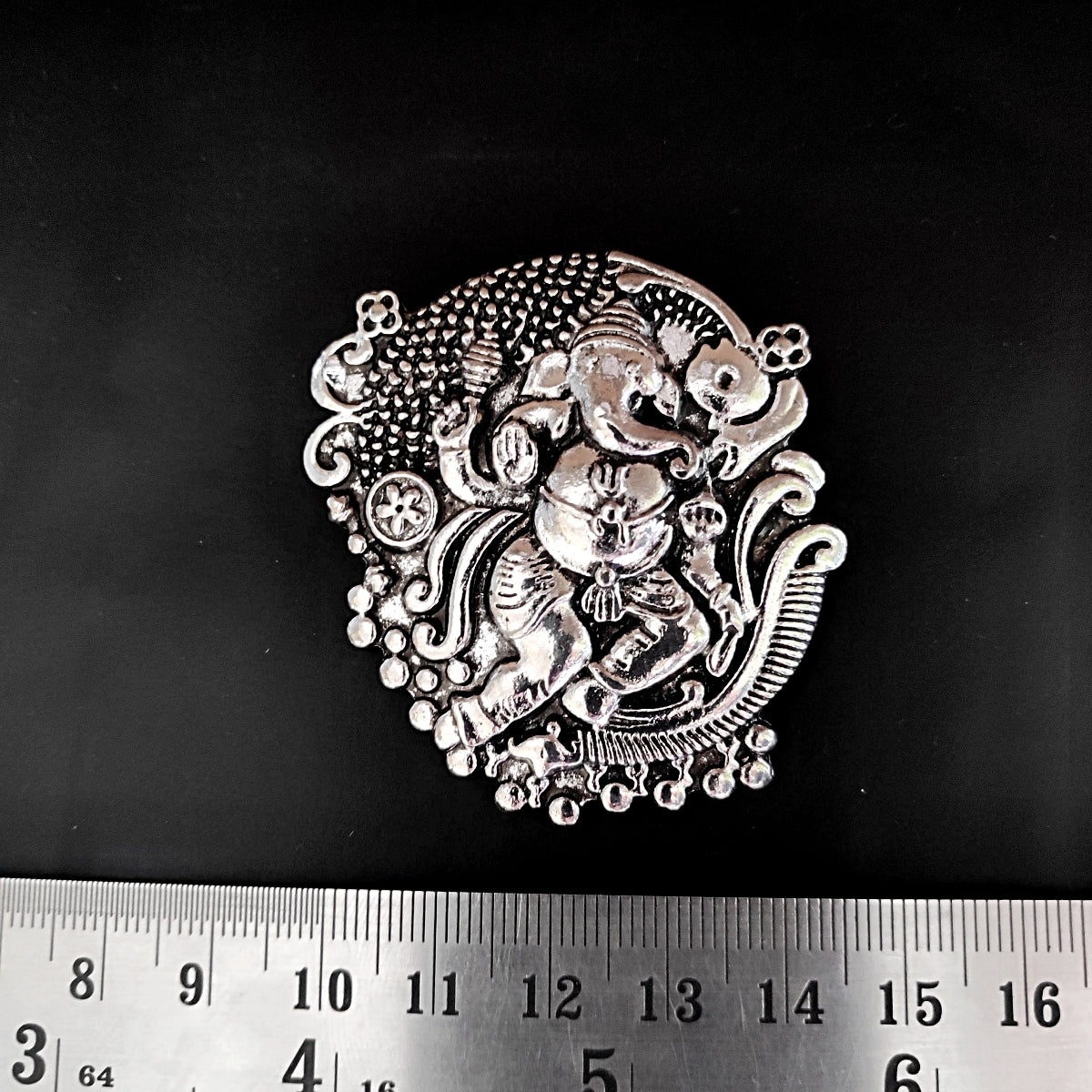 Decorative metal piece with intricate design on a black background, measured against a ruler.