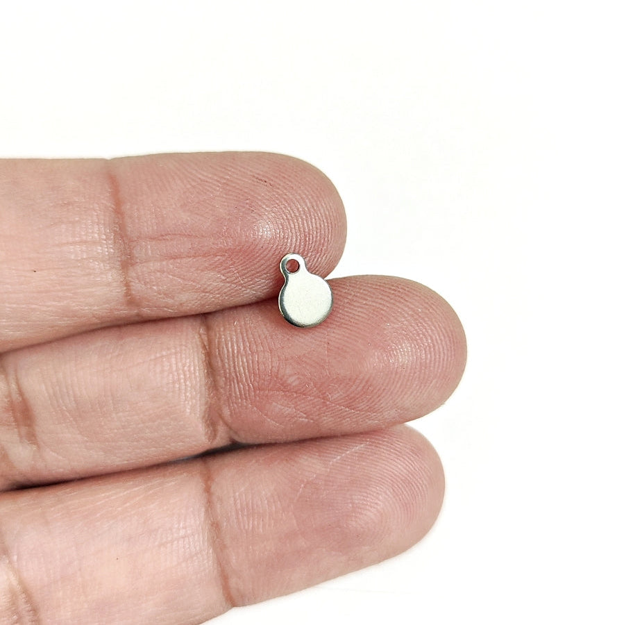 A tiny silver oval charm with a small loop at the top, resting on a person's fingertips against a plain white background.
