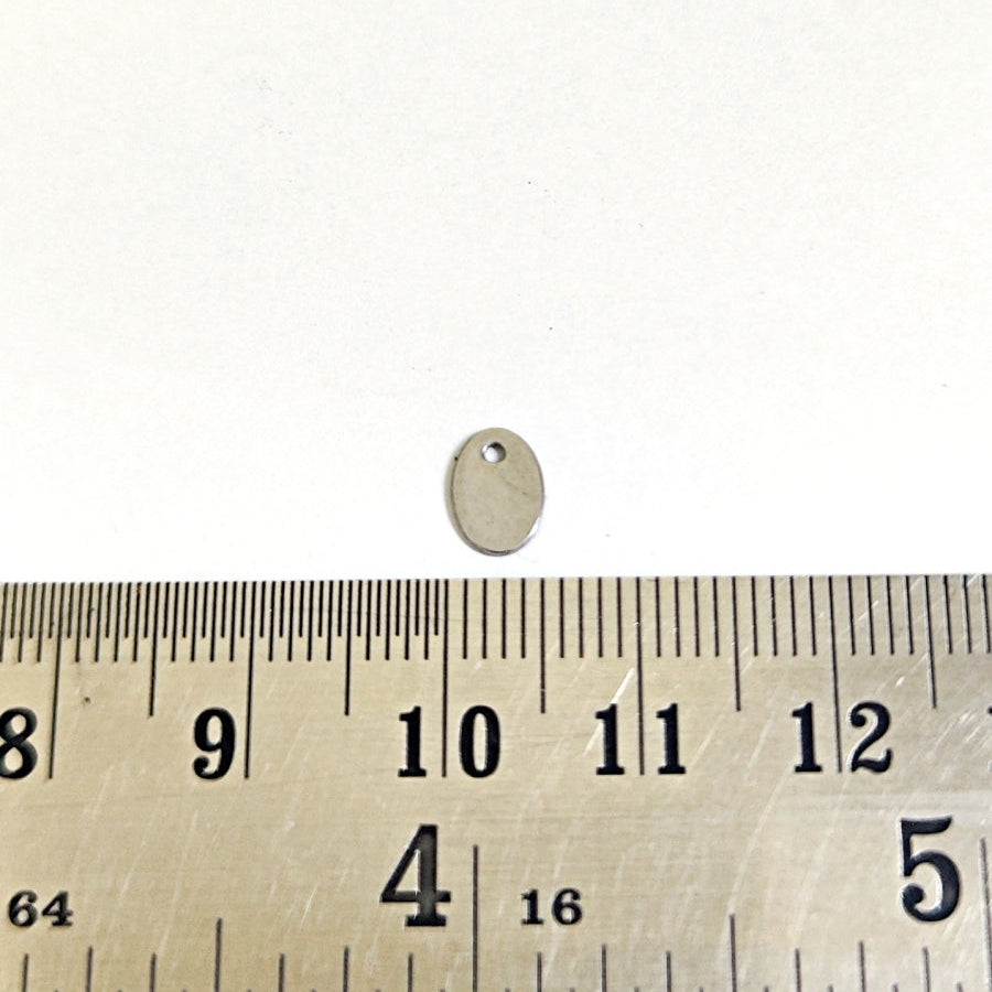 A small oval metal jewelry finding or pendant with a hole, shown above a metal ruler for scale on a white background.
