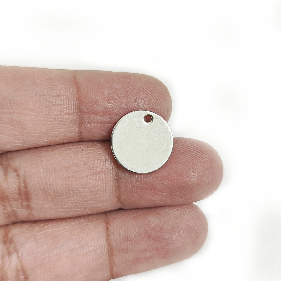 A small blank round metal tag or pendant with a hole, held on a person's fingers to show its size against a white background.