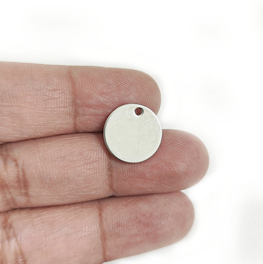 A small blank round metal tag or pendant with a hole, held on a person's fingers to show its size against a white background.