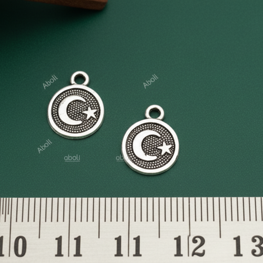 Two silver charms with a moon and star design on a green surface with a ruler for scale.