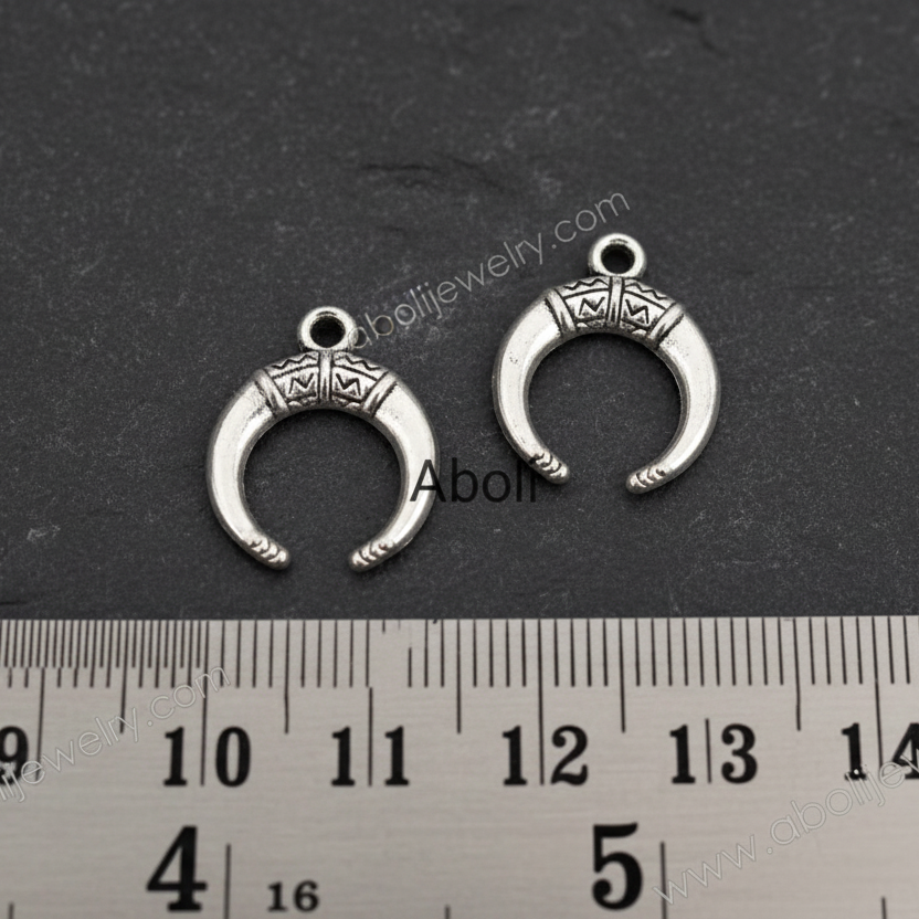 Two silver crescent moon-shaped charms on a dark surface with a ruler for scale.
