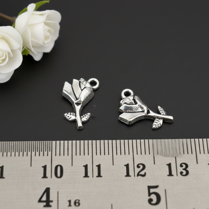 Two silver flower-shaped charms on a black surface with white roses and a ruler for scale.