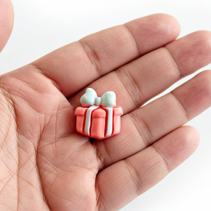 Hand holding small pink gift box-shaped resin cabochon with blue bows on a white background