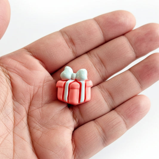Hand holding small pink gift box-shaped resin cabochon with blue bows on a white background